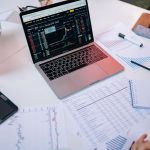 Laptops exibem gráficos do mercado financeiro e documentos sobre uma mesa de trabalho.