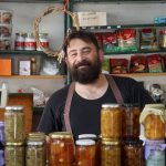 Comerciante atrás de prateleiras com potes de conservas em uma pequena loja.