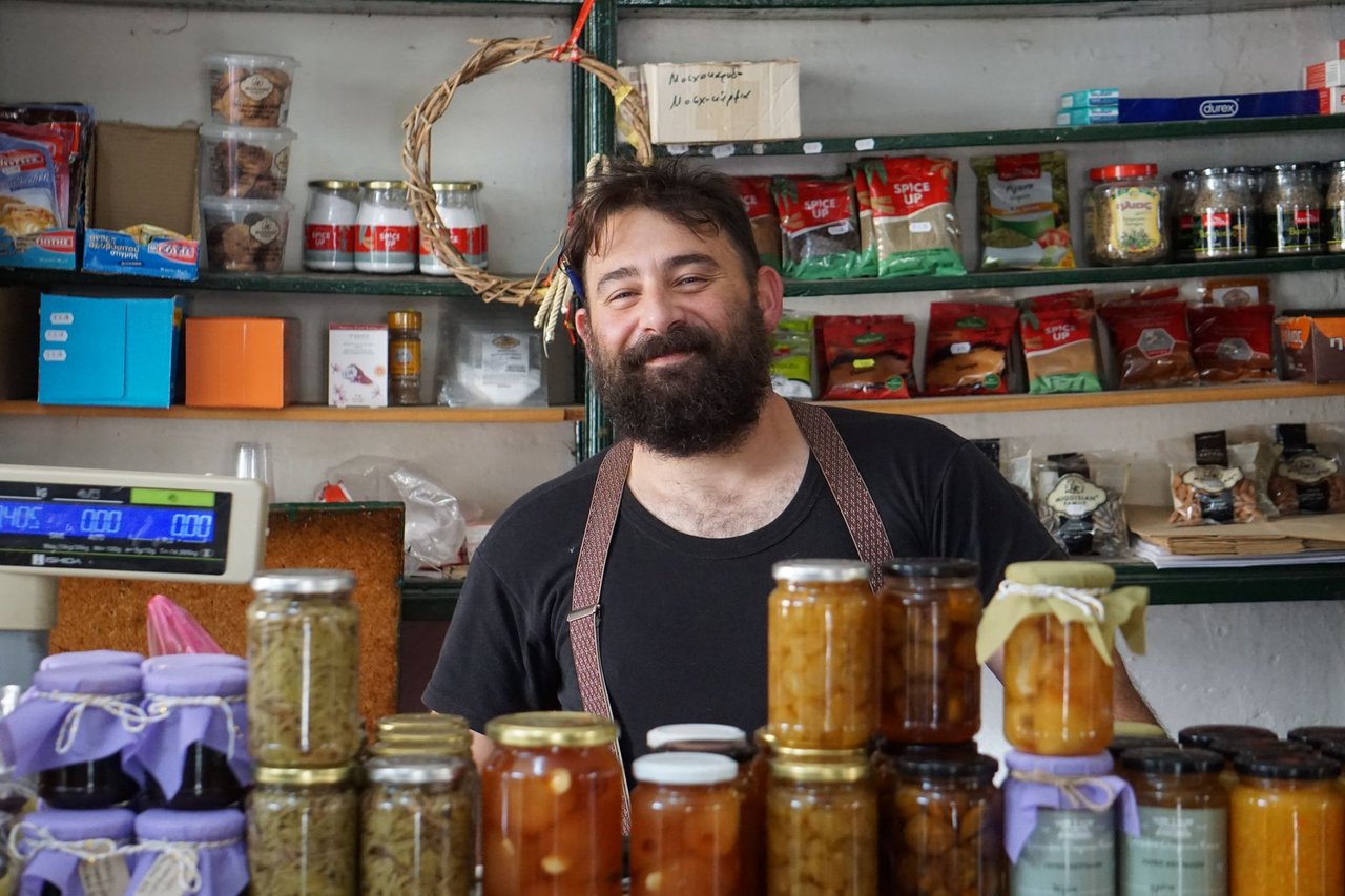 Comerciante atrás de prateleiras com potes de conservas em uma pequena loja.