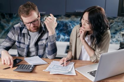Casal analisa contas e documentos ao lado de um laptop enquanto conversa sobre as finanças domésticas.