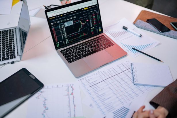 Laptops exibem gráficos do mercado financeiro e documentos sobre uma mesa de trabalho.