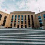 Fachada do Federal Reserve em Washington, vista de baixo, com bandeiras dos Estados Unidos e uma escadaria sob céu nublado.