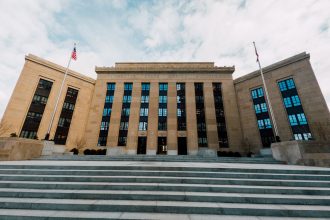 Fachada do Federal Reserve em Washington, vista de baixo, com bandeiras dos Estados Unidos e uma escadaria sob céu nublado.
