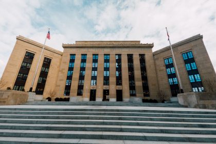 Fachada do Federal Reserve em Washington, vista de baixo, com bandeiras dos Estados Unidos e uma escadaria sob céu nublado.