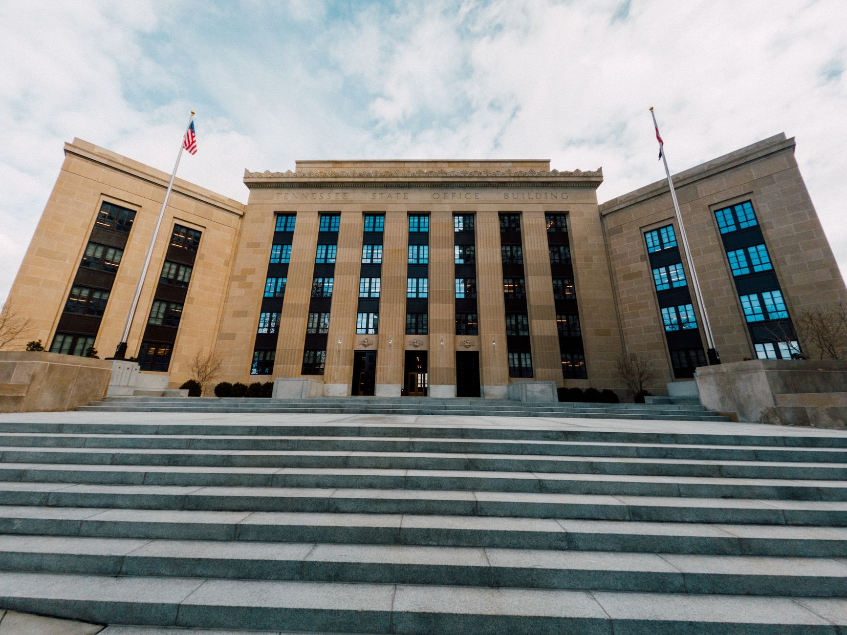 Fachada do Federal Reserve em Washington, vista de baixo, com bandeiras dos Estados Unidos e uma escadaria sob céu nublado.